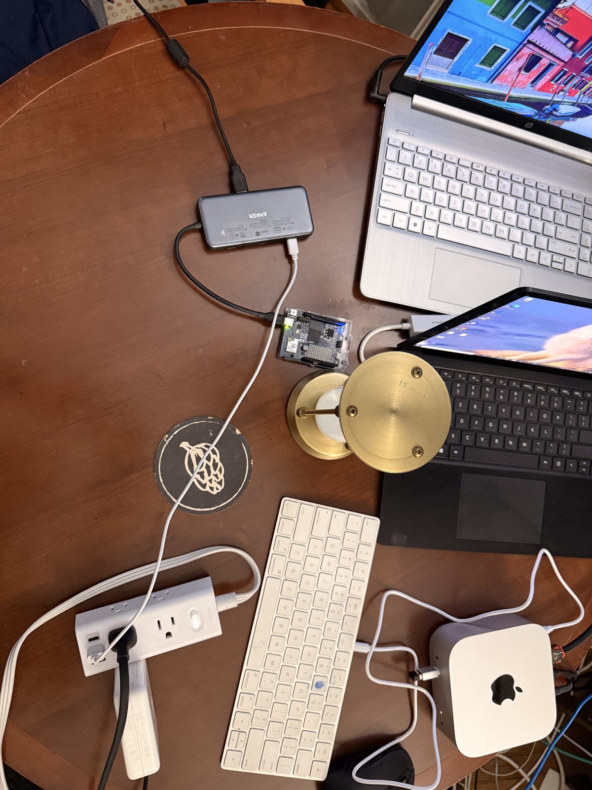 A round wooden table covered in hardware: an Arduino UNO Q board on a USB hub in the center, a Mac mini at bottom right, an Apple keyboard, a power strip, and two laptops